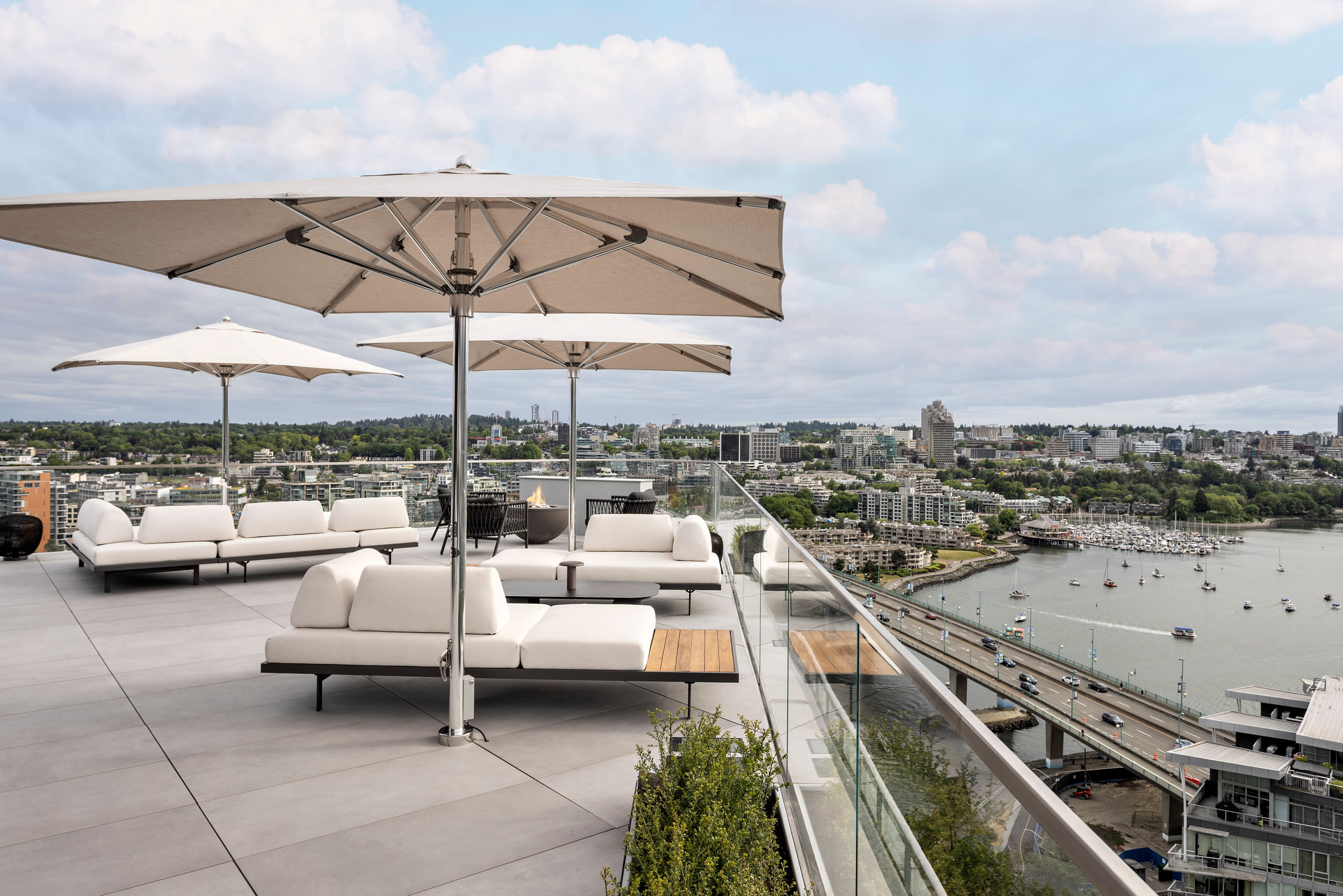 Rooftop outdoor lounge with white sectional couches under large beige umbrellas, overlooking a city harbor.